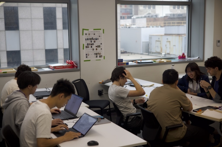 Tisch Summer High School Game Design students sit at tables in a classroom, some working on laptop computers, others working in a group.